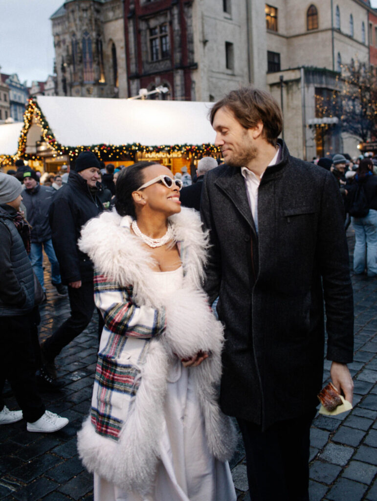 A couple holding hands at a festive European holiday market, smiling at each other while walking together—capturing a cozy, joyful moment during engagement season.