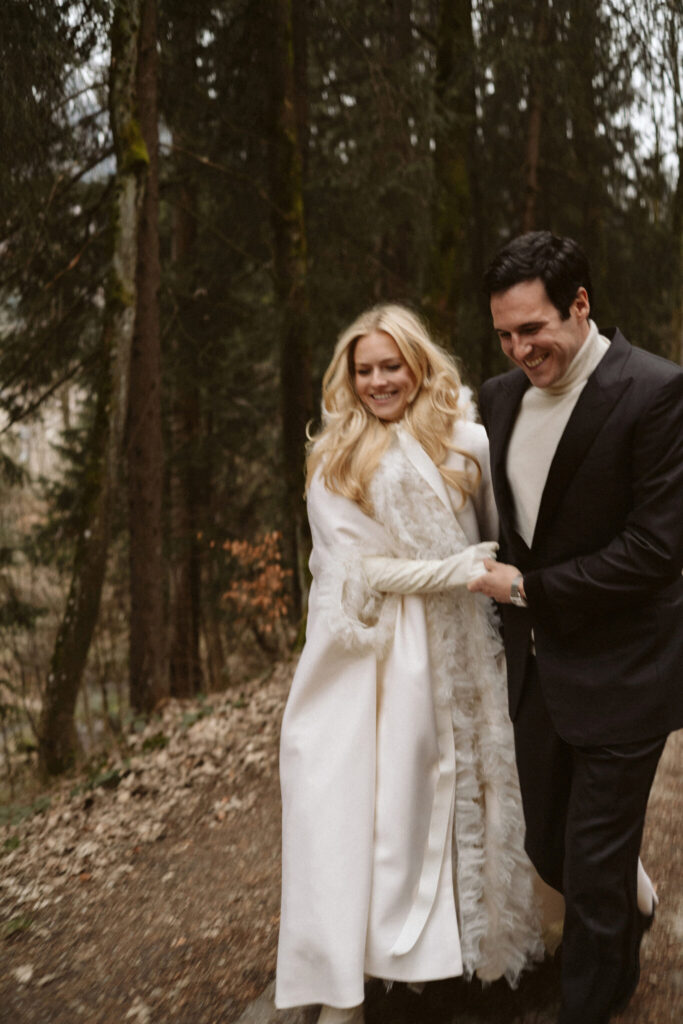 A newly engaged couple walking together through a winter forest, laughing and holding hands—capturing holiday joy during wedding planning season.