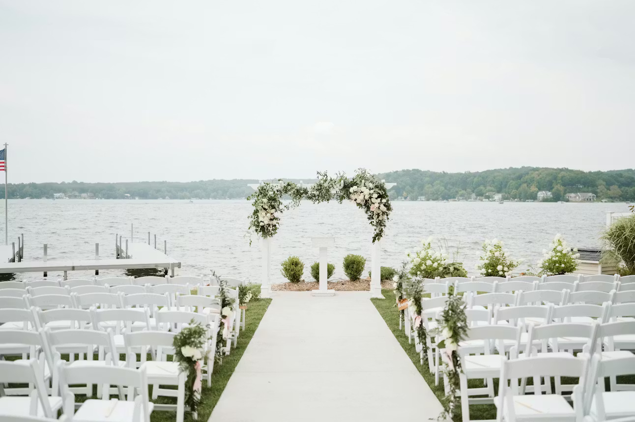 Outdoor waterfront wedding ceremony at Bay Pointe in Southwest Michigan featuring a white floral arch, lakeside views, and elegant white guest seating