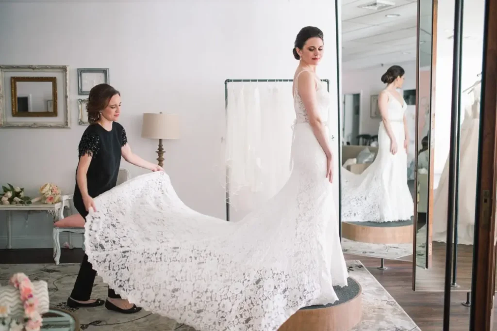 Bride trying on a lace wedding gown in a bridal boutique while an assistant adjusts the train during a wedding dress fitting.