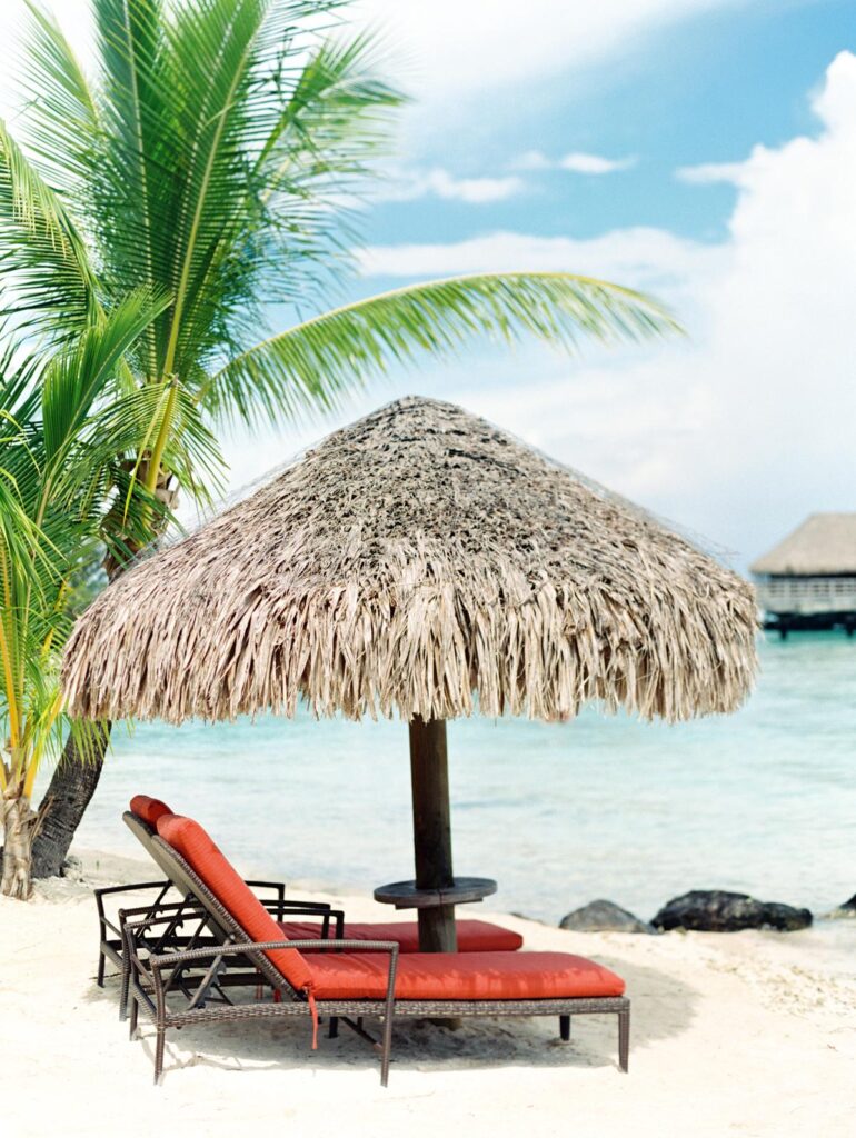 Tropical beach scene with palm tree, thatched umbrella, and lounge chairs overlooking turquoise water at a destination wedding location.