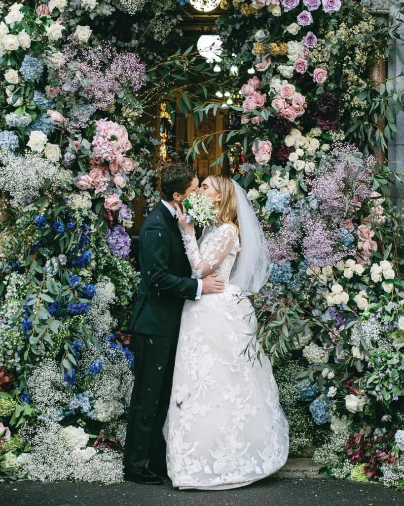 Bride and groom kissing under lush floral ceremony arch with pastel flowers and greenery