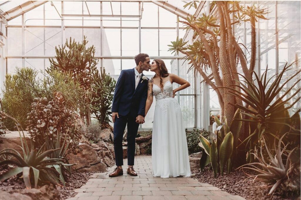 Bride and groom sharing a kiss inside a greenhouse surrounded by lush desert plants and natural light.
