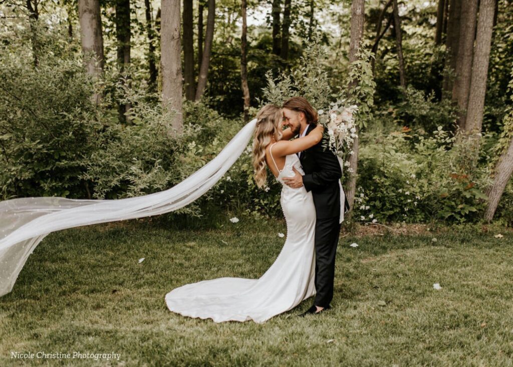Bride and groom embracing outdoors during a West Michigan wedding at Black River Barn with a flowing veil and wooded backdrop.