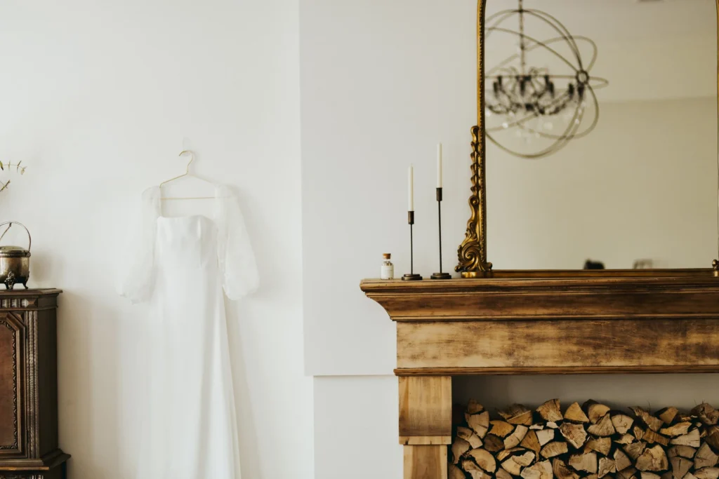 Elegant wedding dress hanging in a bright room beside a rustic fireplace and vintage mirror before the ceremony.