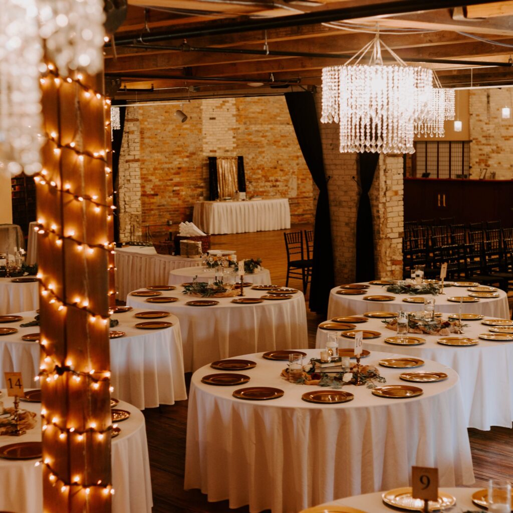 Wedding reception space with round tables, crystal chandeliers, string lights, and exposed brick walls in a historic venue.